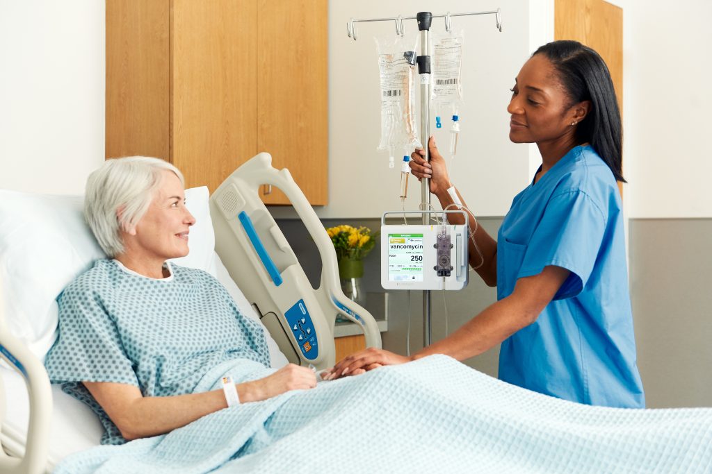 A smiling nurse adjusting an IV drip using the Ivenix Infusion Pump for a content elderly female patient in a hospital bed, with a screen displaying the medication Vancomycin 250 mL.